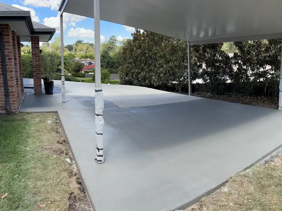 A wide new concrete slab under a carport roof beside a red brick house and lawn