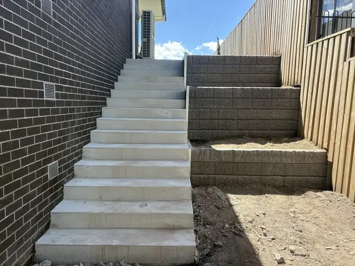 A long flight of new concrete stairs between a dark brick wall and a stepped block retaining wall